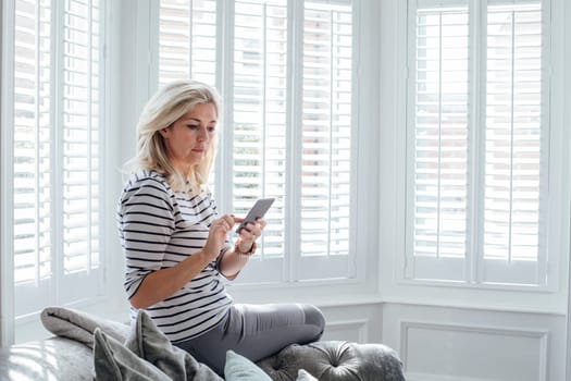 Mature woman using a smart phone. She is sitting on the sofa in her living room.