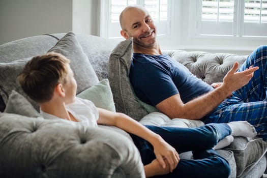 Father and son sitting on the sofa in their home together in teh morning. They are talking and laughing about something they are watching on television.