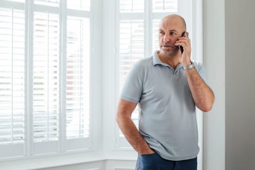 Man talking on the phone. He is standing by the window in his home with a serious expression.