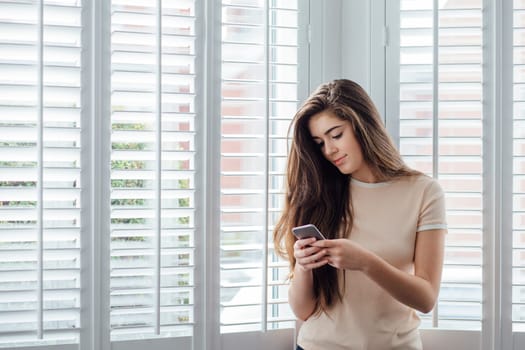 Teenage girl standing by the window in her home, using a smartphone to text.