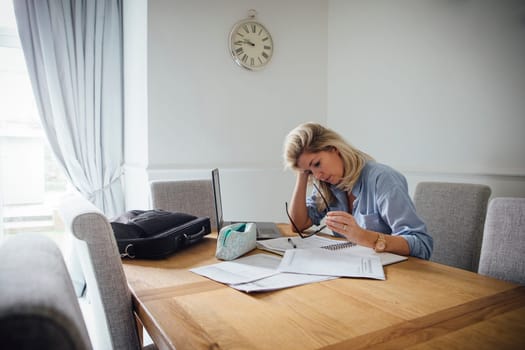 Mature woman working from home. She is sitting at the dining table with a laptop and paperwork.