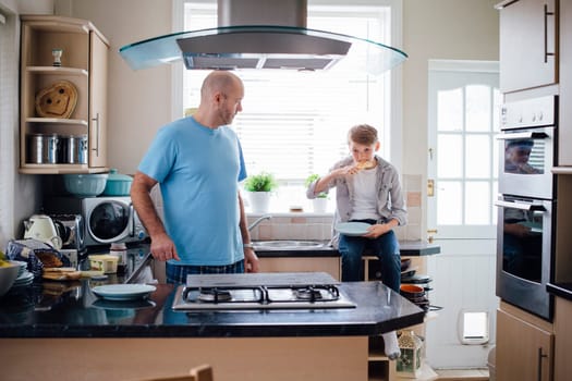 Father and son at home in the kitchen. The boy is sitting on the worktop, eating some toast that his father has made for him.