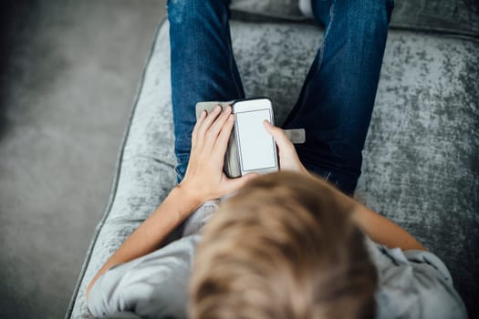 Shot from above of a boy sitting on his sofa at home, using a smartphone.
