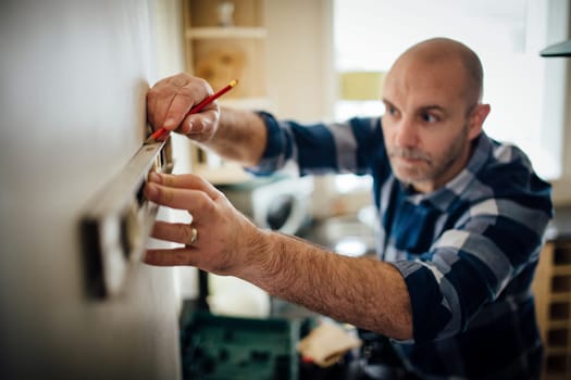 Mature man using a spirit level and marking the wall with a pencil in his kitchen.