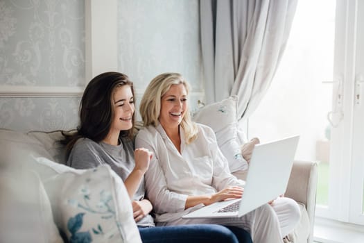Mother and her daughter video calling someone on their laptop. They are smiling and waving at the screen.