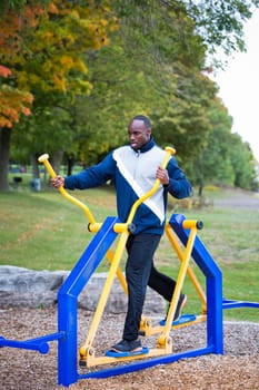 Active black man in training suit exercising on elliptical trainer machine at outdoor gym