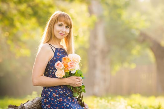 Beautiful young woman looking with a bouquet of roses in hands on a background of green sunny blurred