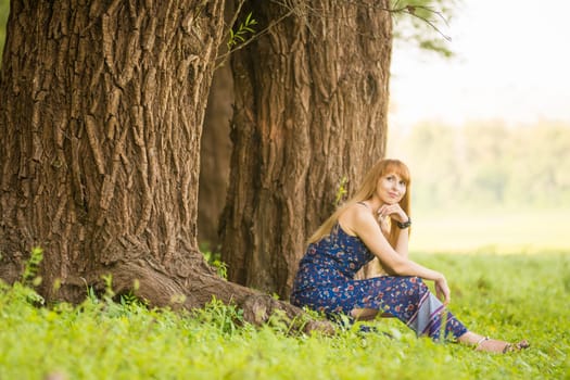 Beautiful young woman sat on the roots of the old large tree and looked up dreamily