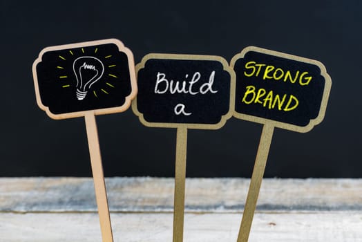 Concept message BUILD A STRONG BRAND and light bulb as symbol for idea written with chalk on wooden mini blackboard labels, defocused chalkboard and wood table in background