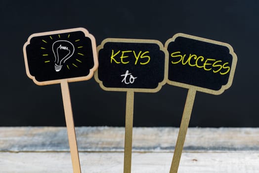 Concept message KEYS TO SUCCESS and light bulb as symbol for idea written with chalk on wooden mini blackboard labels, defocused chalkboard and wood table in background