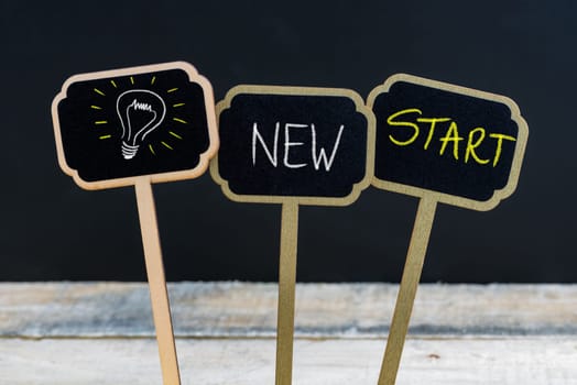 Concept message NEW START and light bulb as symbol for idea written with chalk on wooden mini blackboard labels, defocused chalkboard and wood table in background