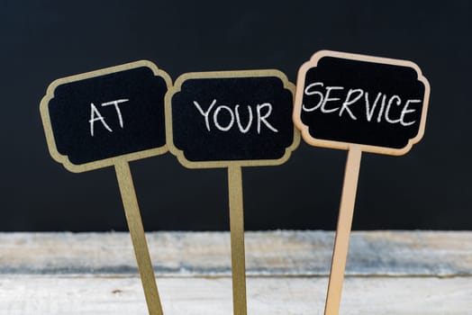 Concept message AT YOUR SERVICE written with chalk on wooden mini blackboard labels, defocused chalkboard and wood table in background