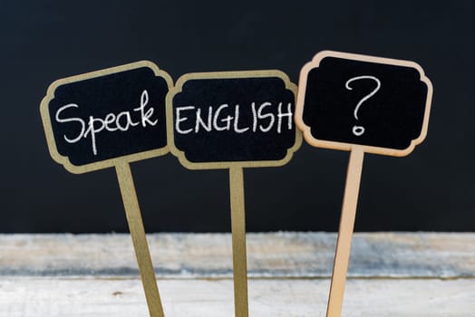 Concept message SPEAK ENGLISH ? written with chalk on wooden mini blackboard labels, defocused chalkboard and wood table in background