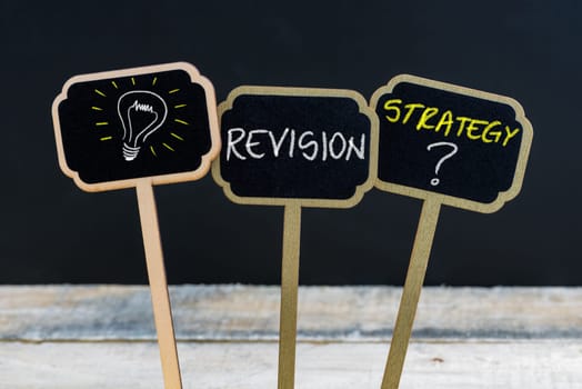 Concept message REVISION STRATEGY ? and light bulb as symbol for idea written with chalk on wooden mini blackboard labels, defocused chalkboard and wood table in background