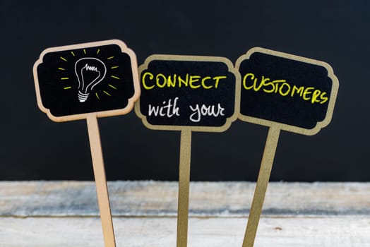 Concept message CONNECT WITH YOUR CUSTOMERS and light bulb as symbol for idea written with chalk on wooden mini blackboard labels, defocused chalkboard and wood table in background