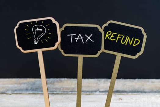 Concept message TAX REFUND and light bulb as symbol for idea written with chalk on wooden mini blackboard labels, defocused chalkboard and wood table in background