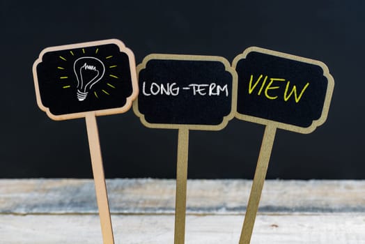 Concept message LONG-TERM VIEW and light bulb as symbol for idea written with chalk on wooden mini blackboard labels, defocused chalkboard and wood table in background