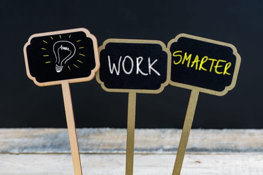 Concept message WORK SMARTER and light bulb as symbol for idea written with chalk on wooden mini blackboard labels, defocused chalkboard and wood table in background