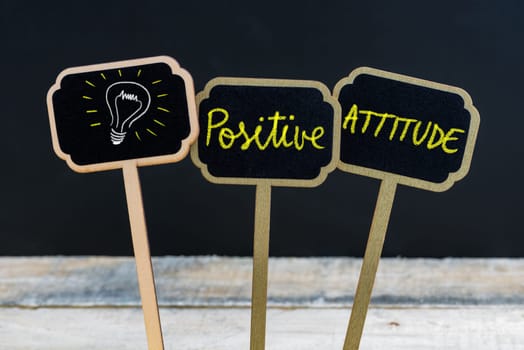 Concept message POSITIVE ATTITUDE and light bulb as symbol for idea written with chalk on wooden mini blackboard labels, defocused chalkboard and wood table in background