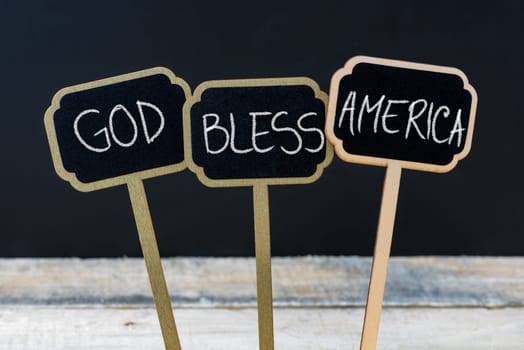Concept message GOD BLESS AMERICA written with chalk on wooden mini blackboard labels, defocused chalkboard and wood table in background