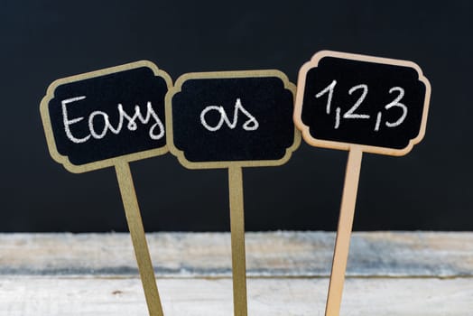 Concept message EASY AS 1,2,3 written with chalk on wooden mini blackboard labels, defocused chalkboard and wood table in background