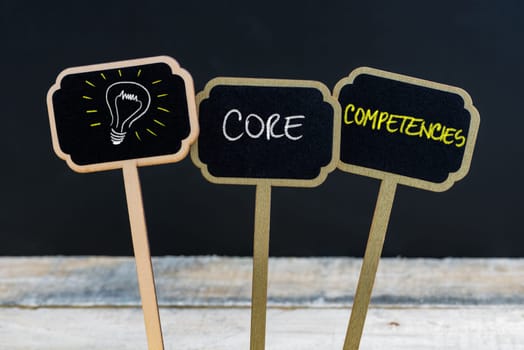 Concept message CORE COMPETENCIES and light bulb as symbol for idea written with chalk on wooden mini blackboard labels, defocused chalkboard and wood table in background