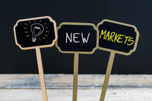 Concept message NEW MARKETS and light bulb as symbol for idea written with chalk on wooden mini blackboard labels, defocused chalkboard and wood table in background