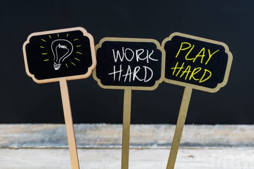 Concept message WORK HARD PLAY HARD and light bulb as symbol for idea written with chalk on wooden mini blackboard labels, defocused chalkboard and wood table in background