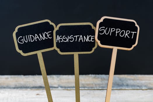 Concept message GUIDANCE, ASSISTANCE, SUPPORT written with chalk on wooden mini blackboard labels, defocused chalkboard and wood table in background