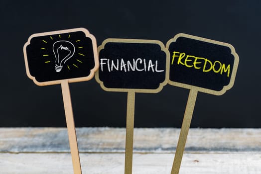 Concept message FINANCIAL FREEDOM and light bulb as symbol for idea written with chalk on wooden mini blackboard labels, defocused chalkboard and wood table in background