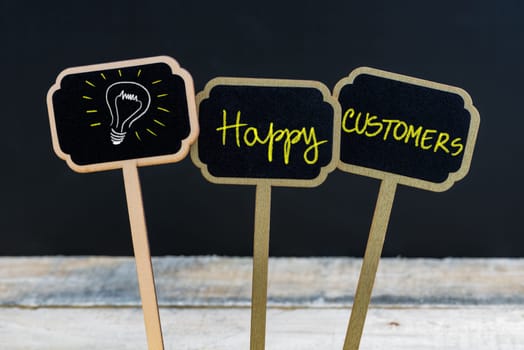 Concept message Happy Customers and light bulb as symbol for idea written with chalk on wooden mini blackboard labels, defocused chalkboard and wood table in background