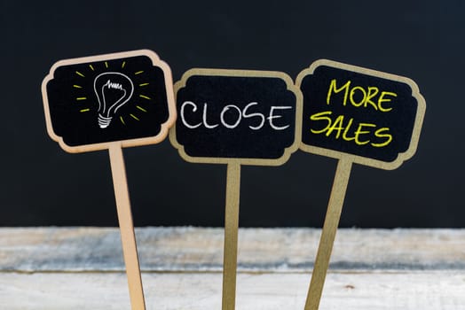 Concept message CLOSE MORE SALES and light bulb as symbol for idea written with chalk on wooden mini blackboard labels, defocused chalkboard and wood table in background