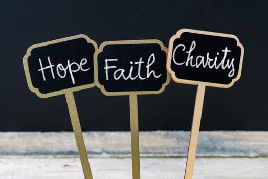 Concept message Hope Faith Charity written with chalk on wooden mini blackboard labels, defocused chalkboard and wood table in background