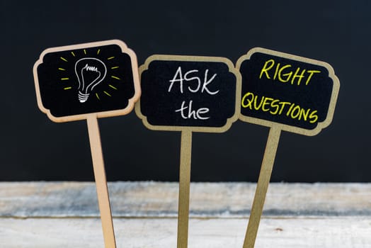 Concept message ASK THE RIGHT QUESTIONS and light bulb as symbol for idea written with chalk on wooden mini blackboard labels, defocused chalkboard and wood table in background