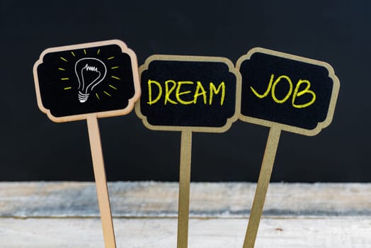 Concept message DREAM JOB and light bulb as symbol for idea written with chalk on wooden mini blackboard labels, defocused chalkboard and wood table in background