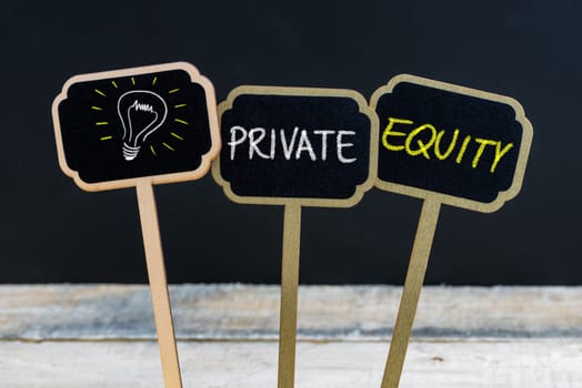 Concept message PRIVATE EQUITY and light bulb as symbol for idea written with chalk on wooden mini blackboard labels, defocused chalkboard and wood table in background