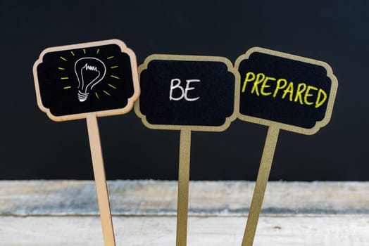 Concept message BE PREPARED and light bulb as symbol for idea written with chalk on wooden mini blackboard labels, defocused chalkboard and wood table in background