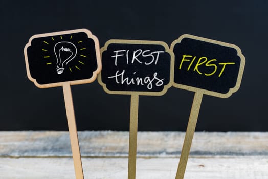 Concept message FIRST THINGS FIRST and light bulb as symbol for idea written with chalk on wooden mini blackboard labels, defocused chalkboard and wood table in background