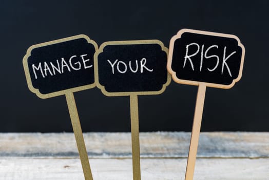 Concept message MANAGE YOUR RISK written with chalk on wooden mini blackboard labels, defocused chalkboard and wood table in background
