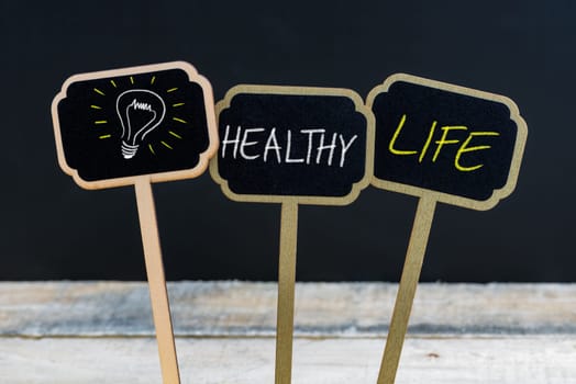 Concept message HEALTHY LIFE and light bulb as symbol for idea written with chalk on wooden mini blackboard labels, defocused chalkboard and wood table in background