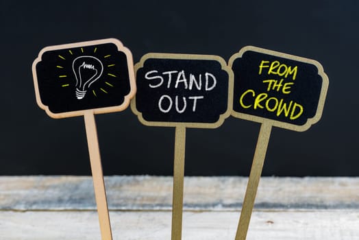 Concept message STAND OUT FROM THE CROWD and light bulb as symbol for idea written with chalk on wooden mini blackboard labels, defocused chalkboard and wood table in background