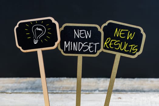 Concept message NEW MINDSET NEW RESULTS and light bulb as symbol for idea written with chalk on wooden mini blackboard labels, defocused chalkboard and wood table in background