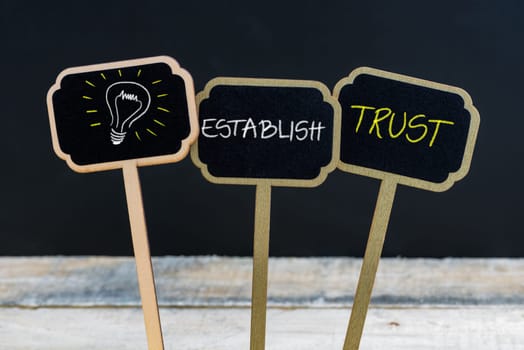 Concept message ESTABLISH TRUST and light bulb as symbol for idea written with chalk on wooden mini blackboard labels, defocused chalkboard and wood table in background