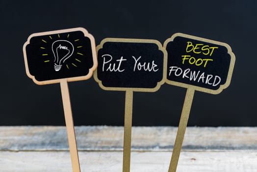 Concept message PUT YOUR BEST FOOT FORWARD and light bulb as symbol for idea written with chalk on wooden mini blackboard labels, defocused chalkboard and wood table in background