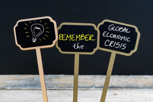 Concept message REMEMBER THE GLOBAL ECONOMIC CRISIS and light bulb as symbol for idea written with chalk on wooden mini blackboard labels, defocused chalkboard and wood table in background