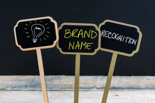 Concept message BRAND NAME RECOGNITION and light bulb as symbol for idea written with chalk on wooden mini blackboard labels, defocused chalkboard and wood table in background