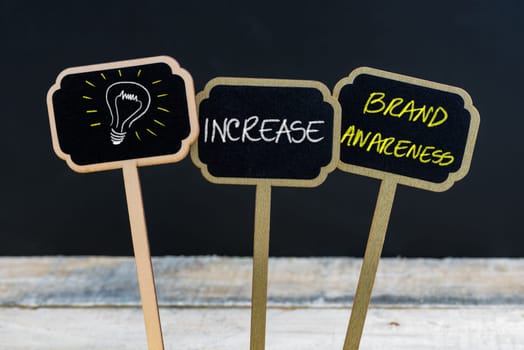 Concept message INCREASE BRAND AWARENESS and light bulb as symbol for idea written with chalk on wooden mini blackboard labels, defocused chalkboard and wood table in background