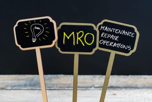 Concept message MRO as Maintenance Repair Operations and light bulb as symbol for idea written with chalk on wooden mini blackboard labels, defocused chalkboard and wood table in background