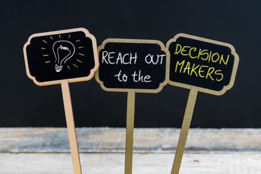 Concept message REACH OUT to the DECISION MAKERS and light bulb as symbol for idea written with chalk on wooden mini blackboard labels, defocused chalkboard and wood table in background