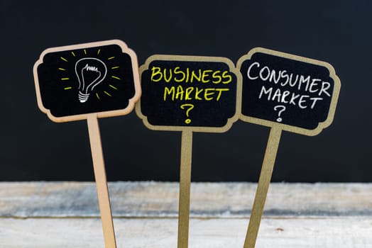 Concept message BUSINESS MARKET versus CONSUMER MARKET and light bulb as symbol for idea written with chalk on wooden mini blackboard labels, defocused chalkboard and wood table in background